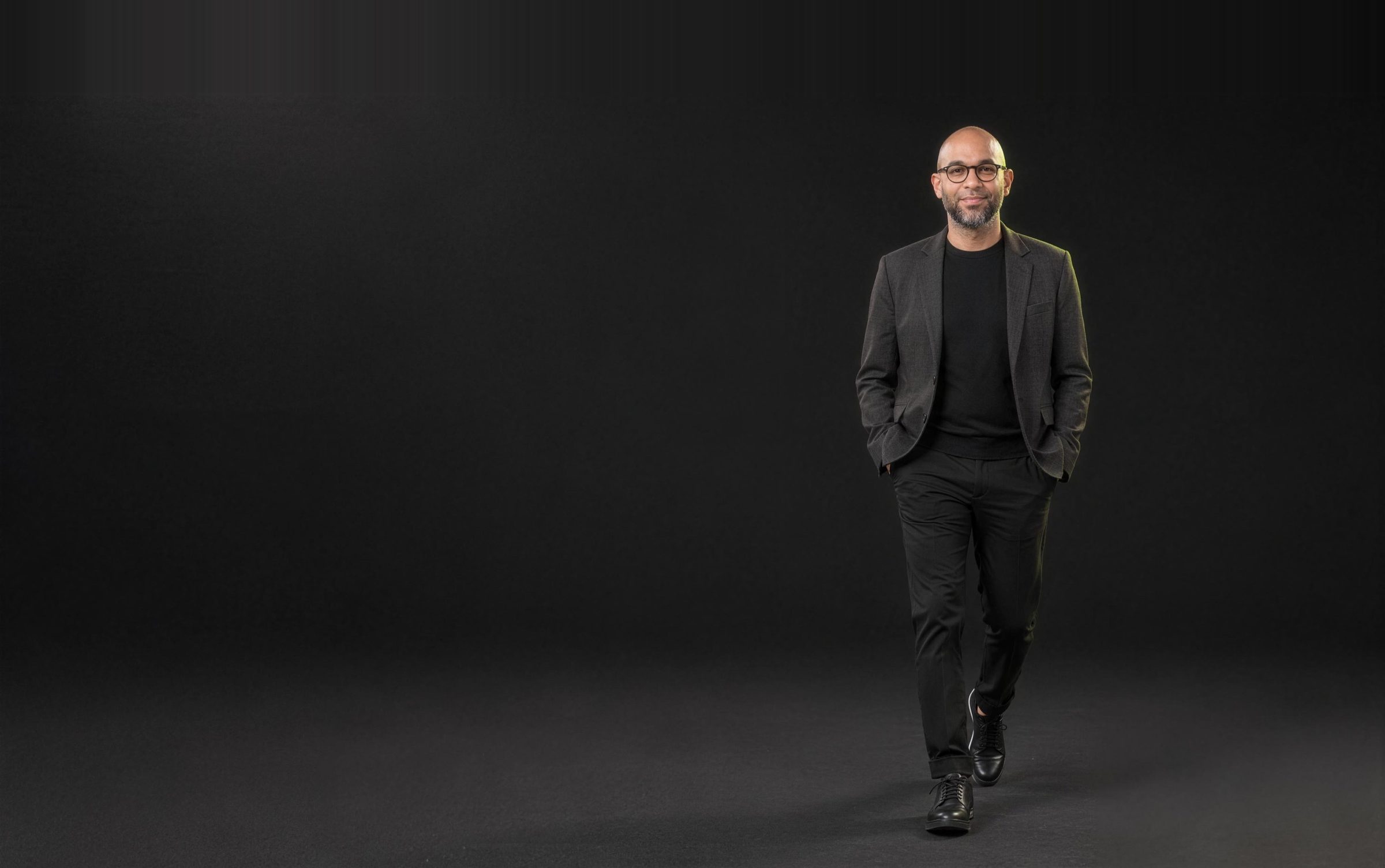 Vikram Ahuja laughing as he walks toward the camera in a dark studio, hands in his pockets